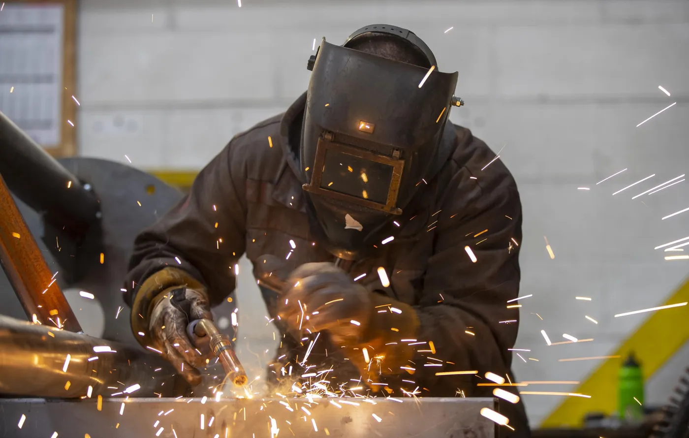 A welder in a full helmet and gauntlets joining steel in the workshop, sparks flying